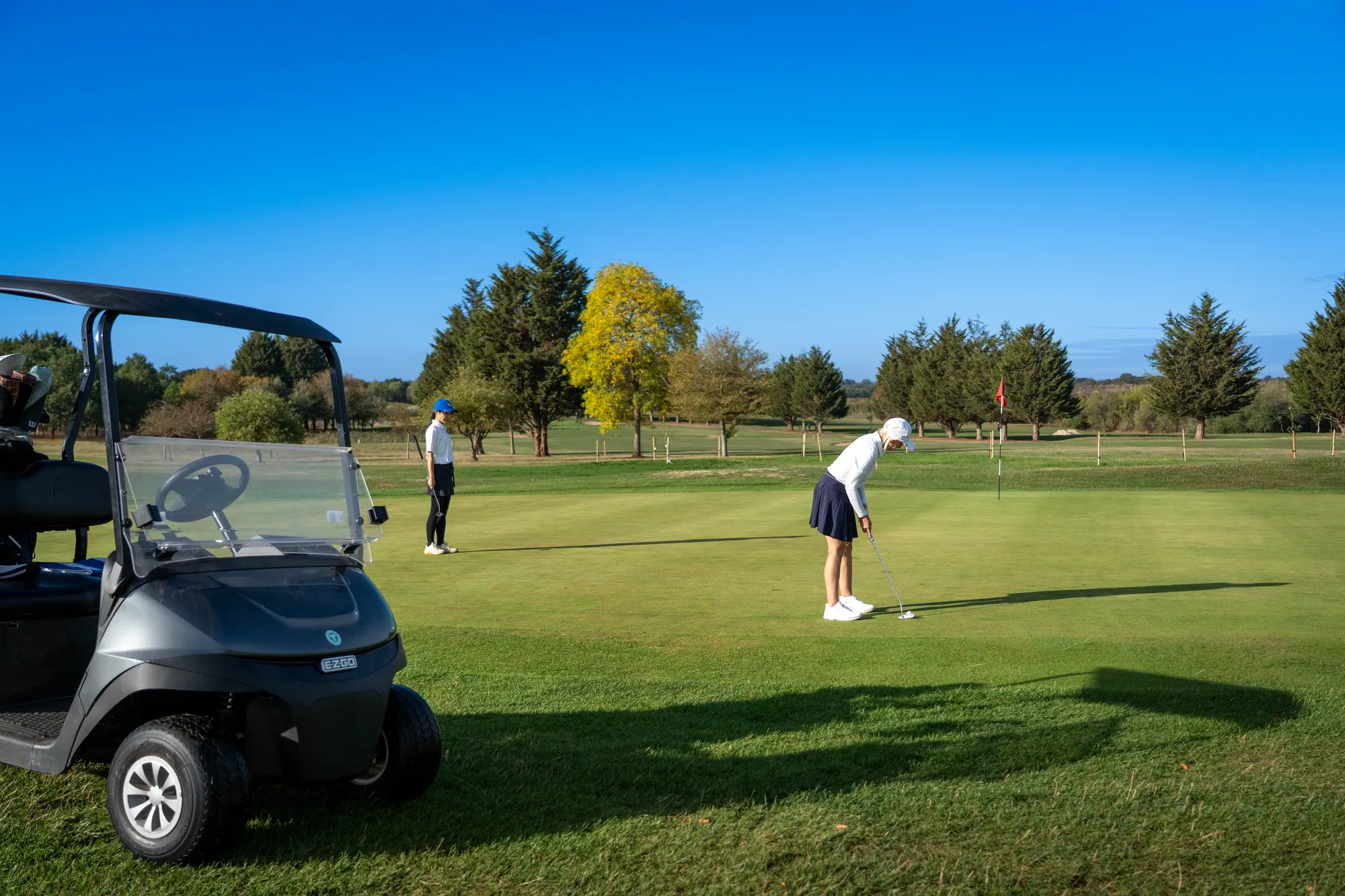 A golfer putting beside a parked buggy on a sunny green