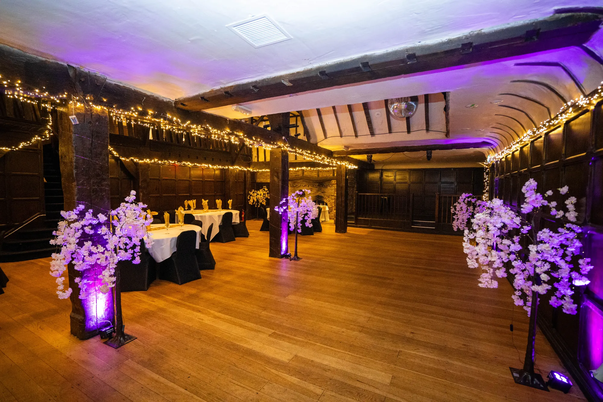 The Baronial Hall set for a wedding, with round tables, purple-lit blossom trees and fairy lights along the beams
