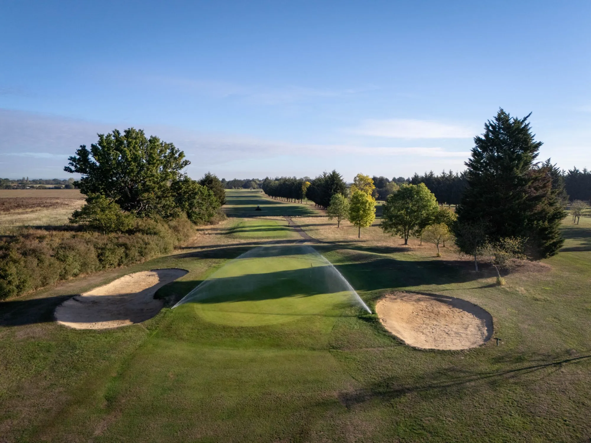 A sprinkler arcing over a green between two bunkers, a tree-lined fairway behind