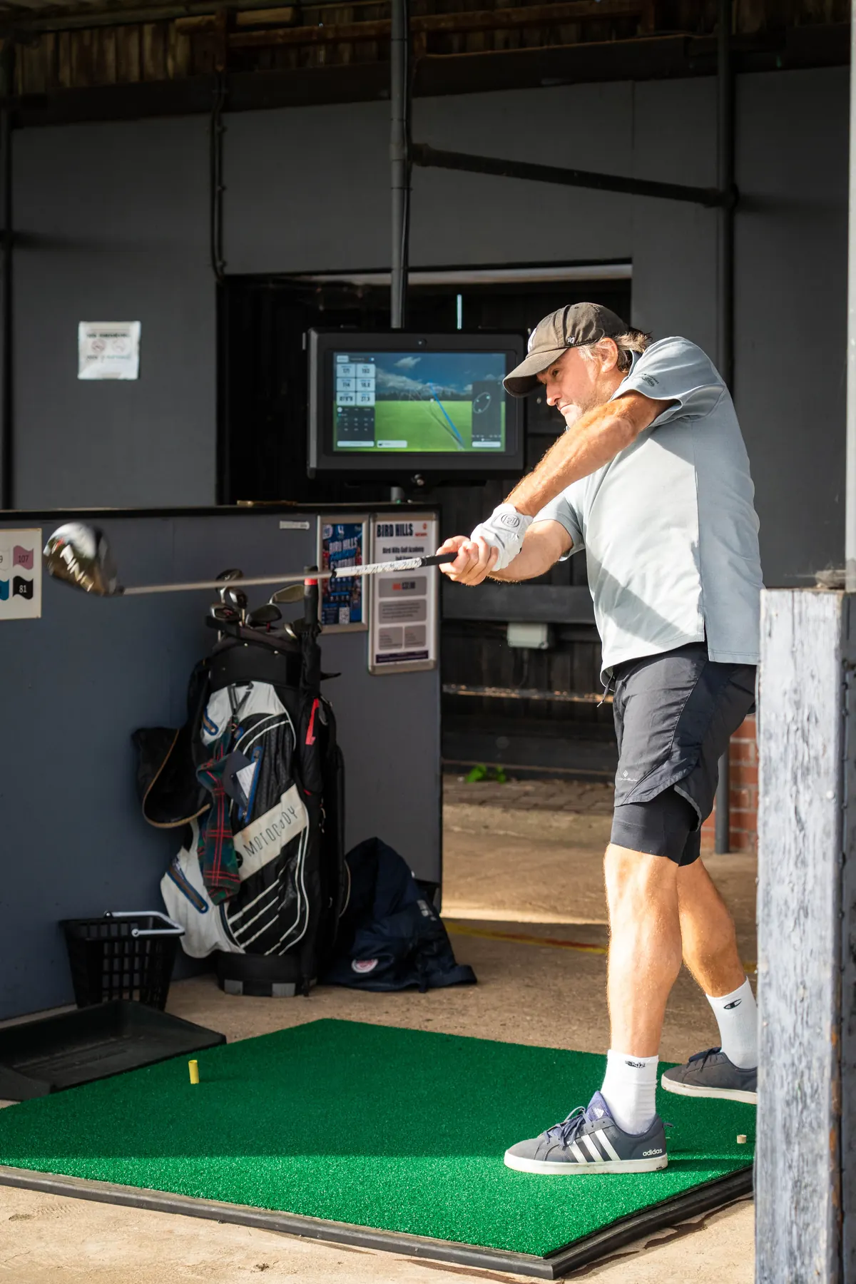 A golfer swinging in a TrackMan bay at the driving range, his shot data on the screen above